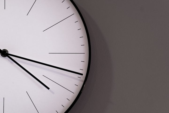 Analoge Uhr mit weißen Ziffernblatt und schwarzen Zeigern an grauer Wand.