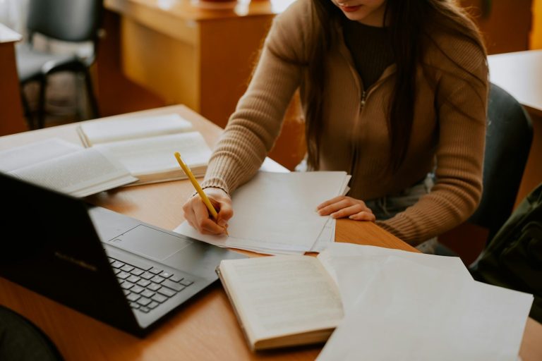 Eine Person schreibt Notizen auf einem Blatt Papier, umgeben von Büchern und einem Laptop.
